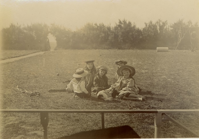 Five children seated on grass all wearing smart clothes and hats. Small dog in centre of group.