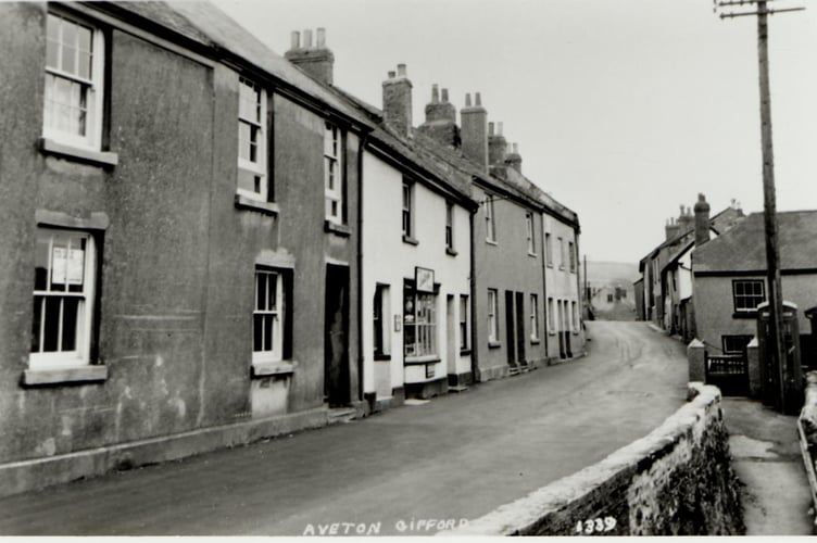 Telephone box on right hand side of Fore Street, Aveton Gifford. Provision store on left. Brick wall in front of telephone box. Around 1910.